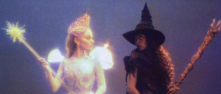 Two contrasting witches stand side by side on a dark stage: one in a glowing white gown with a crown and sparkling wand, the other in a black dress and pointed hat holding a broom, both facing each other dramatically.