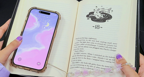A person holding a book as if they are reading but they have their phone in front of the left page. 